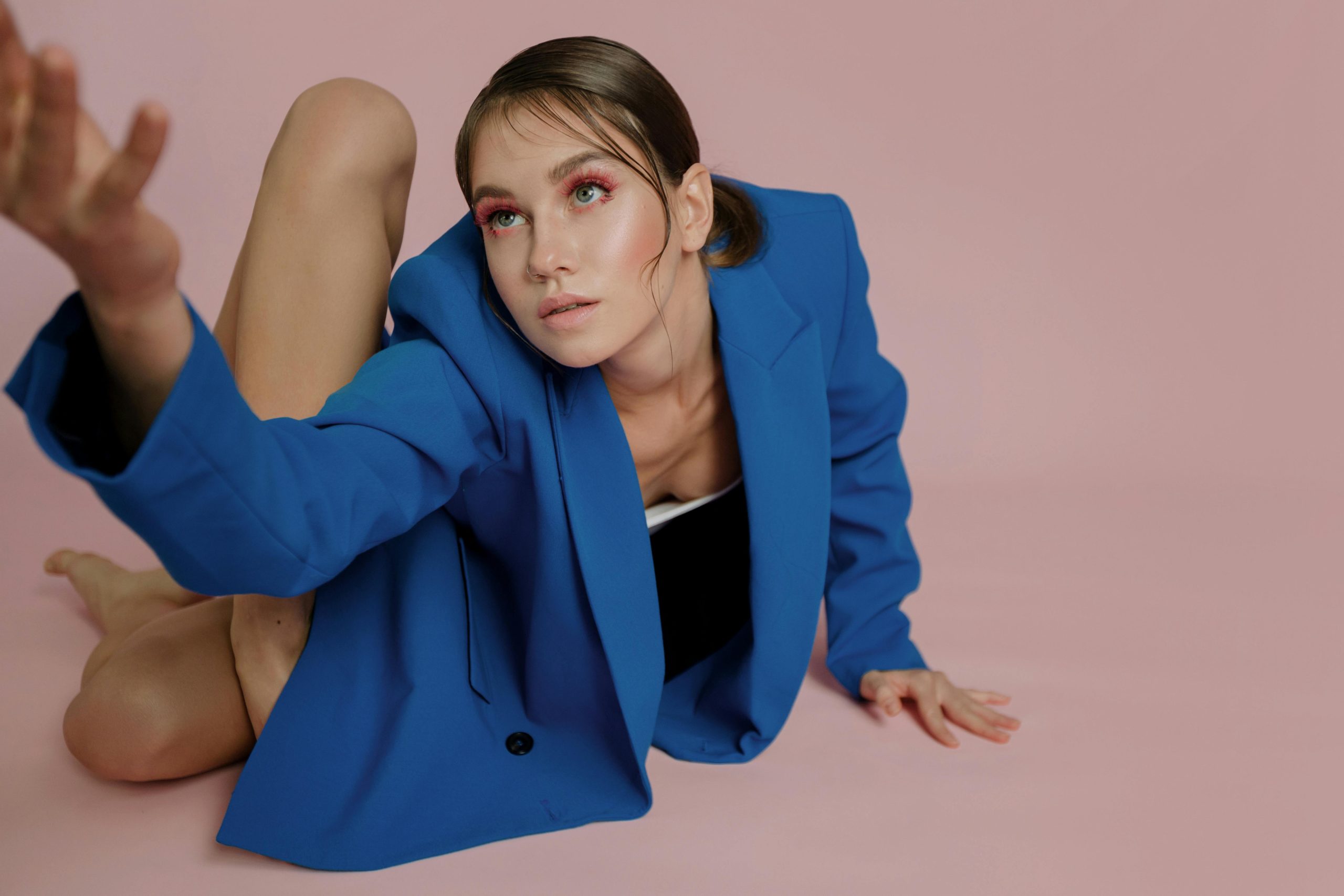 Model posing with a flexible body in a blue jacket against a pink studio background.