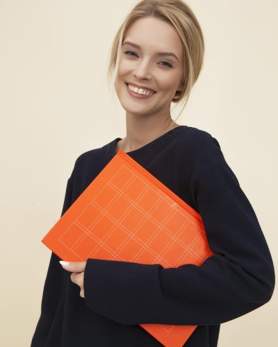 A cheerful woman smiles while holding an orange folder against a neutral background.